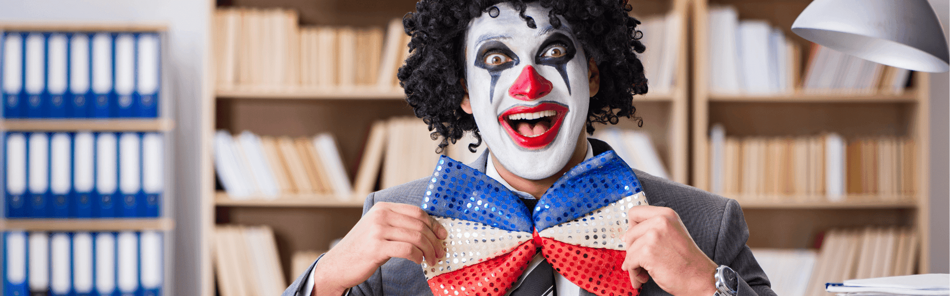 Ein Clown in Business-Anzug mit geschminktem Gesicht und großer rot-weiß-blauer Schleife posiert lächelnd in einem Büro mit Regalen im Hintergrund.