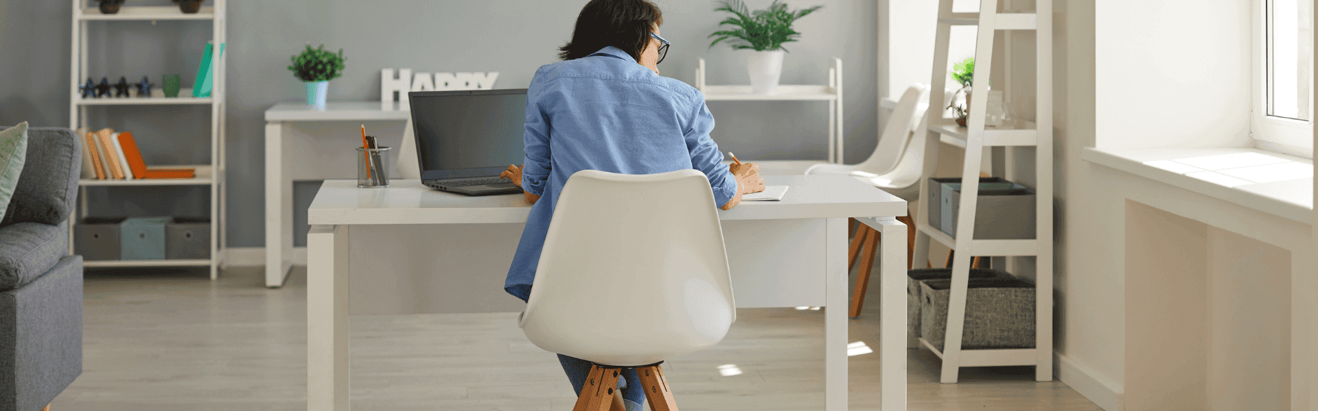 Eine junge Frau arbeitet konzentriert am Laptop an einem Schreibtisch im Homeoffice, umgeben von Unterlagen und einer Kaffeetasse, während Tageslicht durch das Fenster fällt.