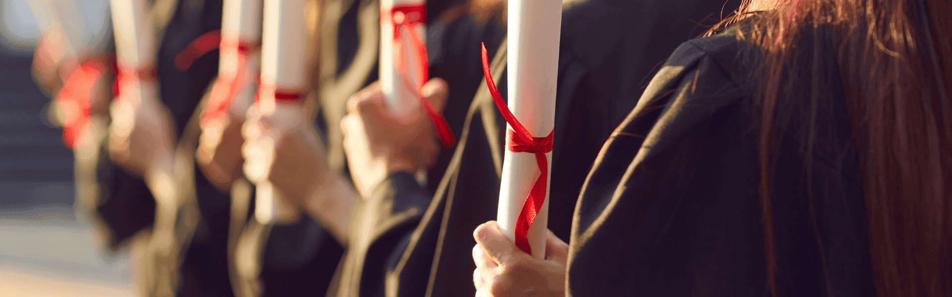Mehrere Absolventen in schwarzen Talaren halten Diplome mit roten Bändern in der Hand, stehen in einer Reihe im Sonnenlicht.